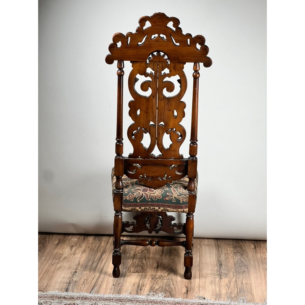 The Antique Carolean Revival Side Chair, c. late 19th century, features an intricately carved tall backrest and armrests, floral fabric upholstery, and sits on a wooden floor against a plain background. Brand: Unknown.
