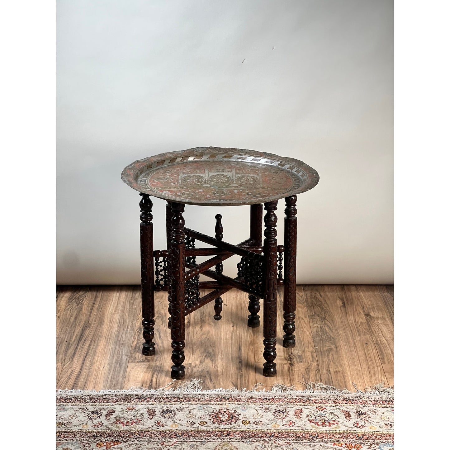 Moroccan or Syrian Moorish folding tray table featuring a hand-chased metal tray top, intricate wooden legs, and an elaborate lattice design, displayed on a hardwood floor near a patterned rug against a plain wall.