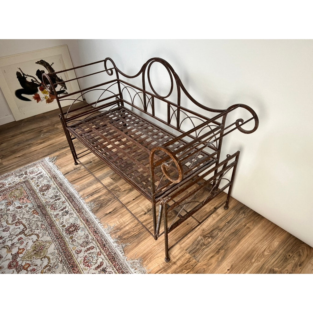 A Vintage Regency Style Wrought Iron Porch Glider with ornate scrollwork evokes a vintage garden feel as it sits on a wooden floor by a patterned rug; framed artwork is propped against the white wall behind it.