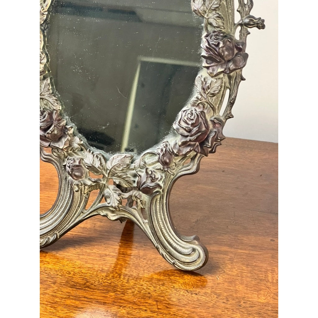 An Antique Art Nouveau Silvered Cast Brass Rose Vanity Dressing Mirror, dated 1909, with an ornate floral frame of detailed roses and leaves, rests on a polished wood surface; only its lower half is shown in the image. Brand: Unknown.
