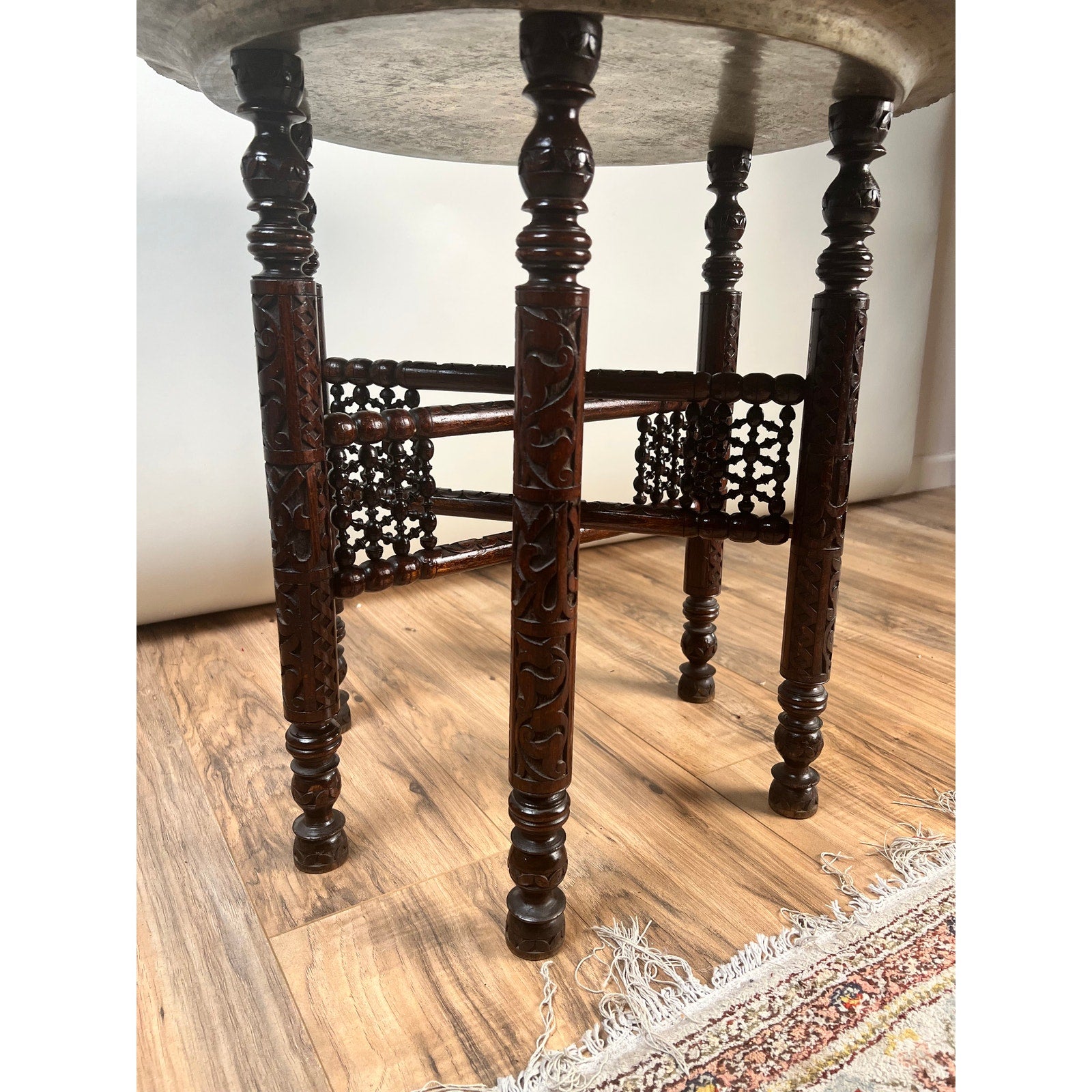 Intricately carved wooden legs support the round tabletop of a Moroccan or Syrian Moorish folding tray table, which sits atop a wood floor beside the edge of a patterned rug.
