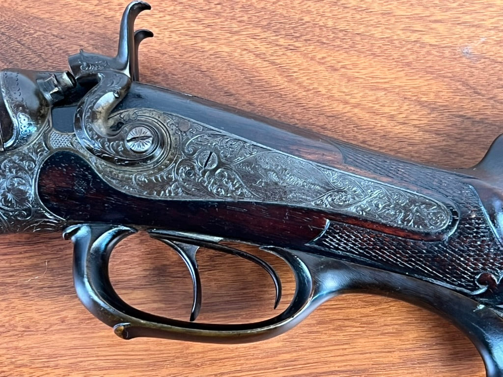 Close-up of an intricately engraved Antique Christoph Funke Suhl German Damascus Double Barrel Hammer Shotgun, c. 1880, featuring ornate metal patterns and a dark carved wooden stock from Pearl City Antiques. Set against a wood grain background, this beautifully crafted piece boasts elegant Damascus barrels that highlight exceptional artistry and meticulous attention to detail.