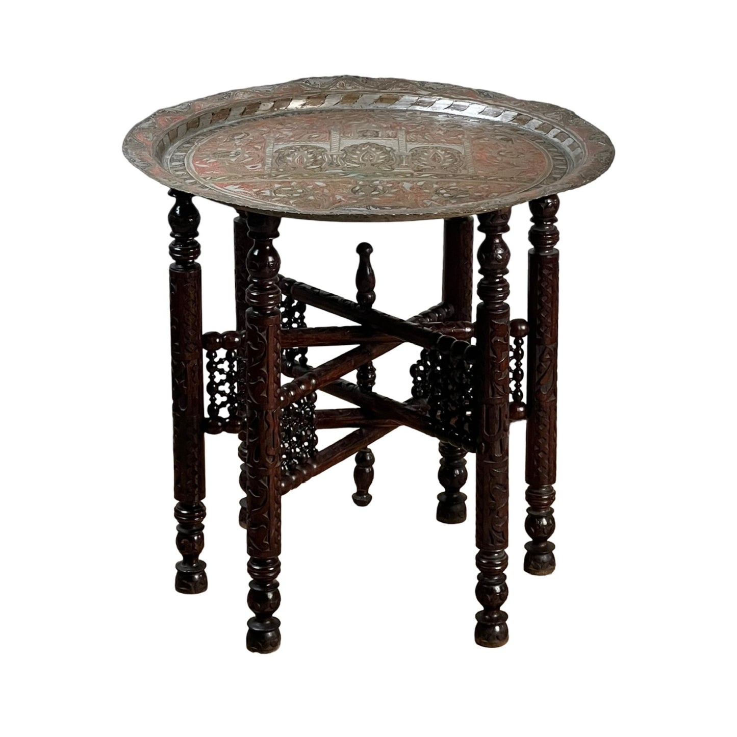 A round, ornate metal tray tops an intricately carved wooden stand with six legs and decorative cross-bracing panels, showcasing the timeless elegance of a Moroccan or Syrian Moorish folding tray table.