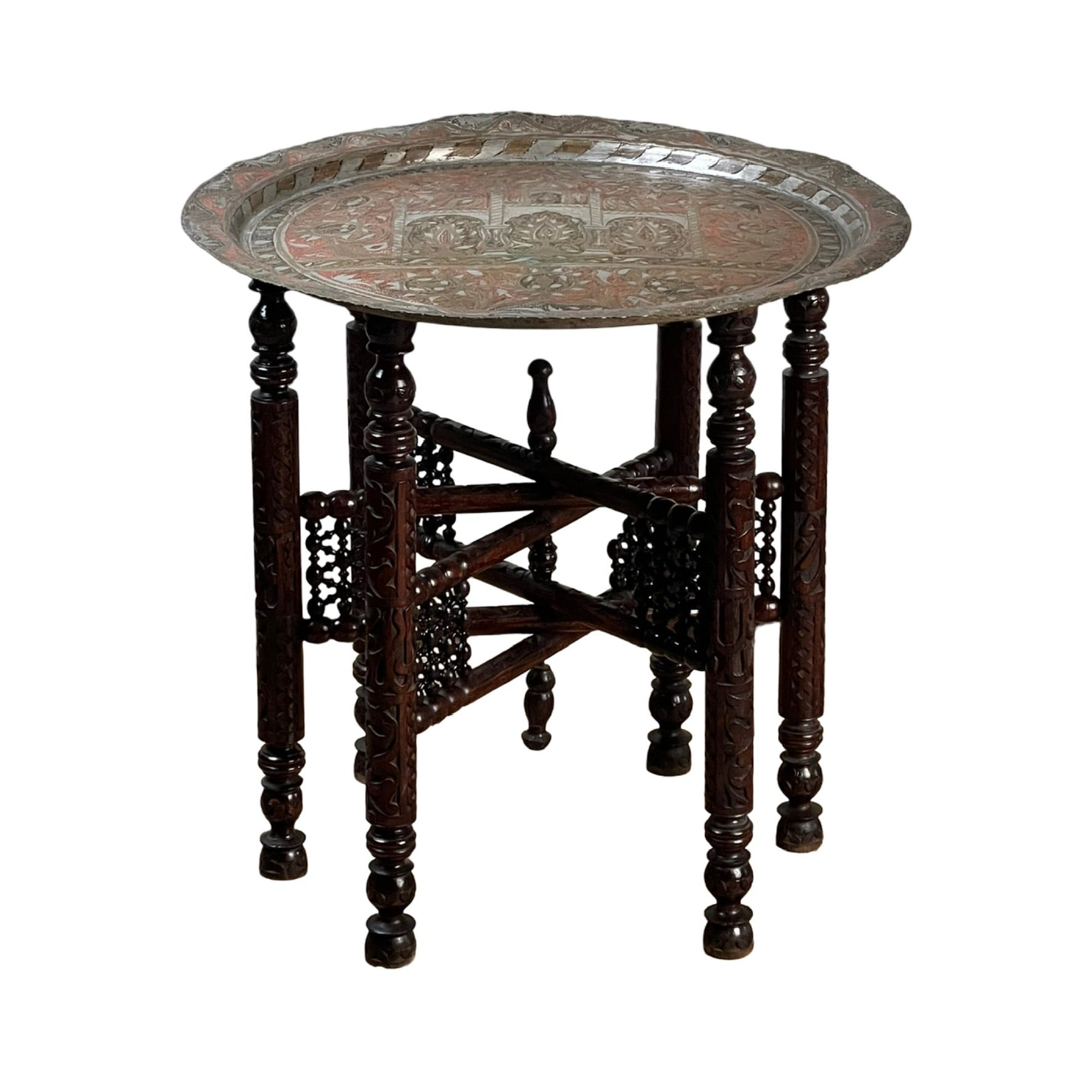 A round, ornate metal tray tops an intricately carved wooden stand with six legs and decorative cross-bracing panels, showcasing the timeless elegance of a Moroccan or Syrian Moorish folding tray table.