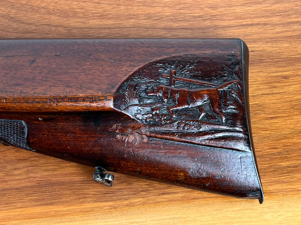Close-up of the carved wooden stock on a c. 1880 Antique Christoph Funke Suhl German Damascus Double Barrel Hammer Shotgun from Pearl City Antiques, featuring detailed engravings of a hunting dog and trees, shown against a wood grain background.