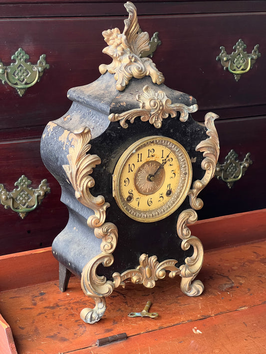 An Antique Cast Iron Rococo Mantle Clock by Ansonia Clock Co. New York from Pearl City Antiques, featuring gold accents and Roman numerals, rests on a wooden surface with a mahogany dresser and brass handles in the background.