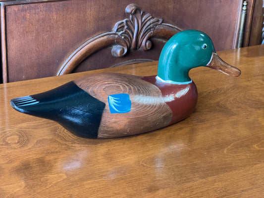 The Pearl City Antiques Hand Carved Duck Decoy, featuring a green head, brown chest, black tail, and blue wing details, sits on a polished wood table in front of an ornate wooden chair—ideal for any decoy collection.
