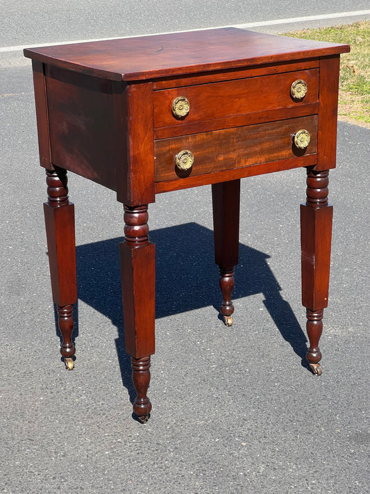 The Antique New York Sheraton Two Drawer Stand by Pearl City Antiques, featuring brass pulls and turned legs on wheels, is displayed outdoors on asphalt, exemplifying early 19th-century design.