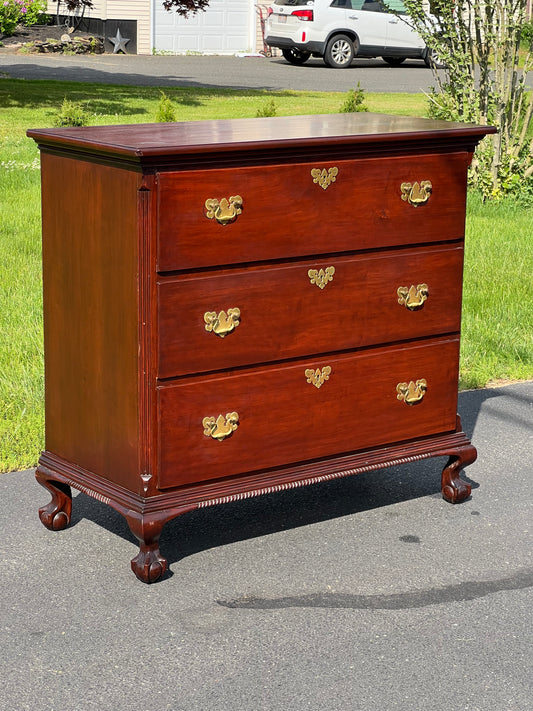 The Antique Mahogany Three Drawer Chippendale Chest c. 1820 by Pearl City Antiques, with brass handles and ornate carved feet, stands on a driveway in sunlight, exemplifying classic Chippendale furniture design.