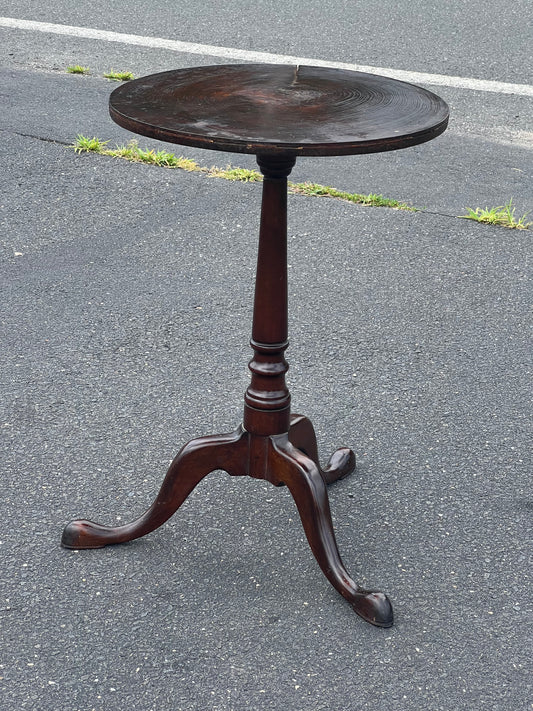 The Antique Queen Anne Snake Foot Candle Stand with Old Growth Cookie Top by Pearl City Antiques, a small round dark wood table with three curved legs, stands on an asphalt surface near a road and some grass.