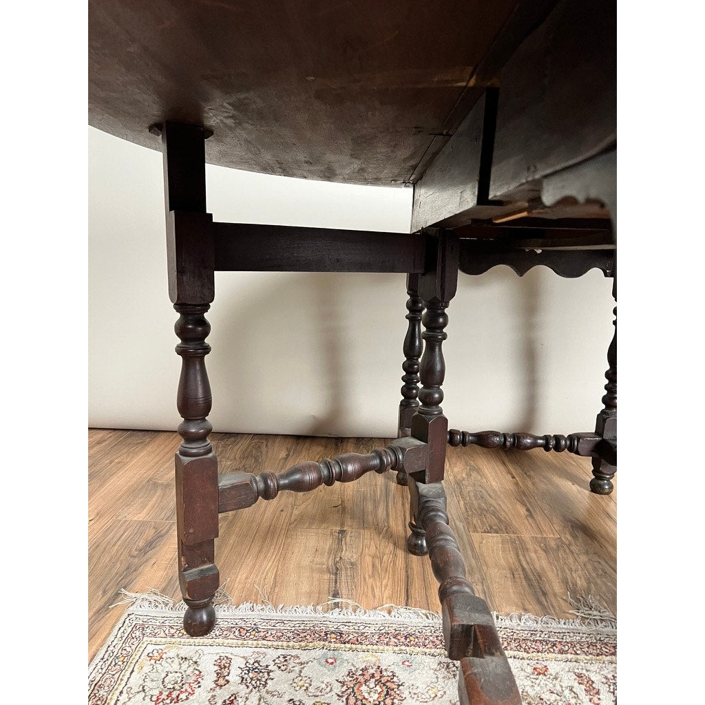 A close-up of the wooden legs and crossbars of an Antique English William & Mary Walnut One Drawer Gateleg Dining Table (early 18th c.) on a wooden floor, with part of a patterned rug seen beneath—a classic period design.