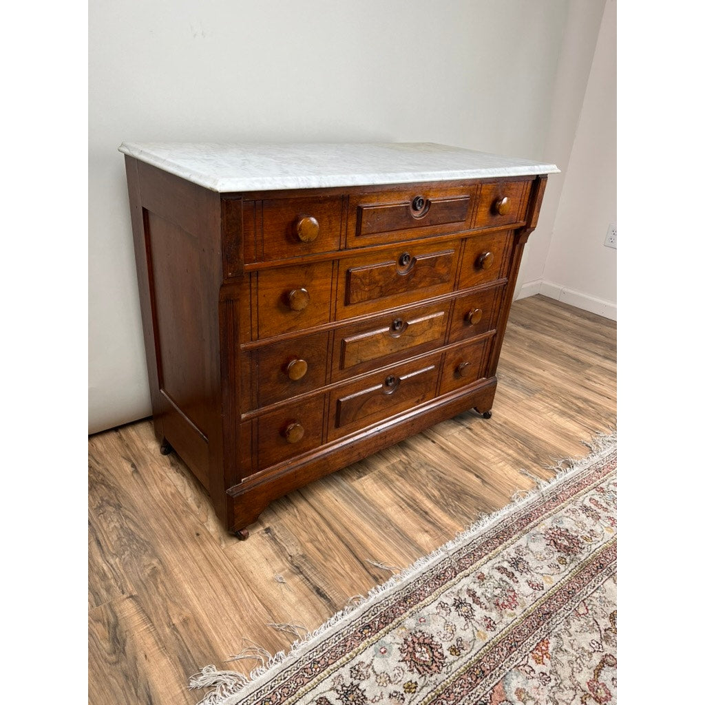 An Antique Victorian Marble Top Four Drawer Chest (c. 1910) stands on a wood floor by a patterned rug, its white marble surface gleaming softly in the gently lit room. Brand: Unknown.