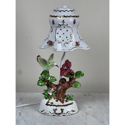 The Pearl City Antiques Vintage Lena Liu Porcelain Hummingbird Accent Lamp features a delicately painted hummingbird hovering near a vibrant red hibiscus flower on a tree-stump base. This porcelain table lamp includes a bell-shaped shade adorned with intricate floral patterns, lattice cutouts, and gold trim set against a light background, creating an elegant vintage-style floral accent.