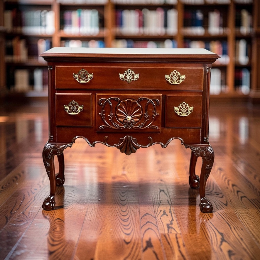A Vintage Solid Mahogany Chippendale Style Lowboy by Councill Craftsmen with brass handles stands on a polished wood floor in a library, bookshelves of books blurred in the background.