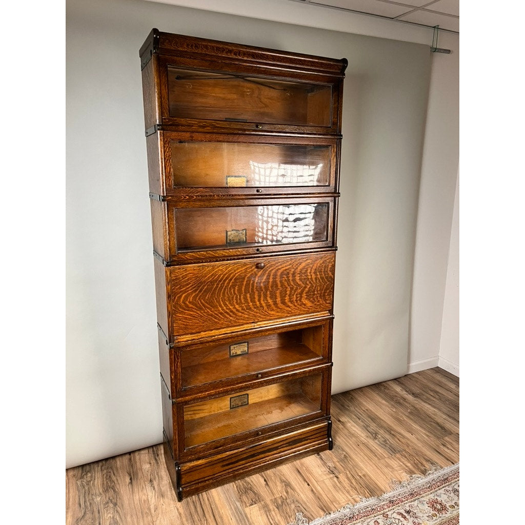 Antique Globe Wernicke Quarter Sawn Oak 6-Stack Barrister Bookcase Secretary Desk (c. 1905–1915), featuring six glass-front stacking sections, brass label holders, and dark trim, is set on a wood floor against a light wall background.