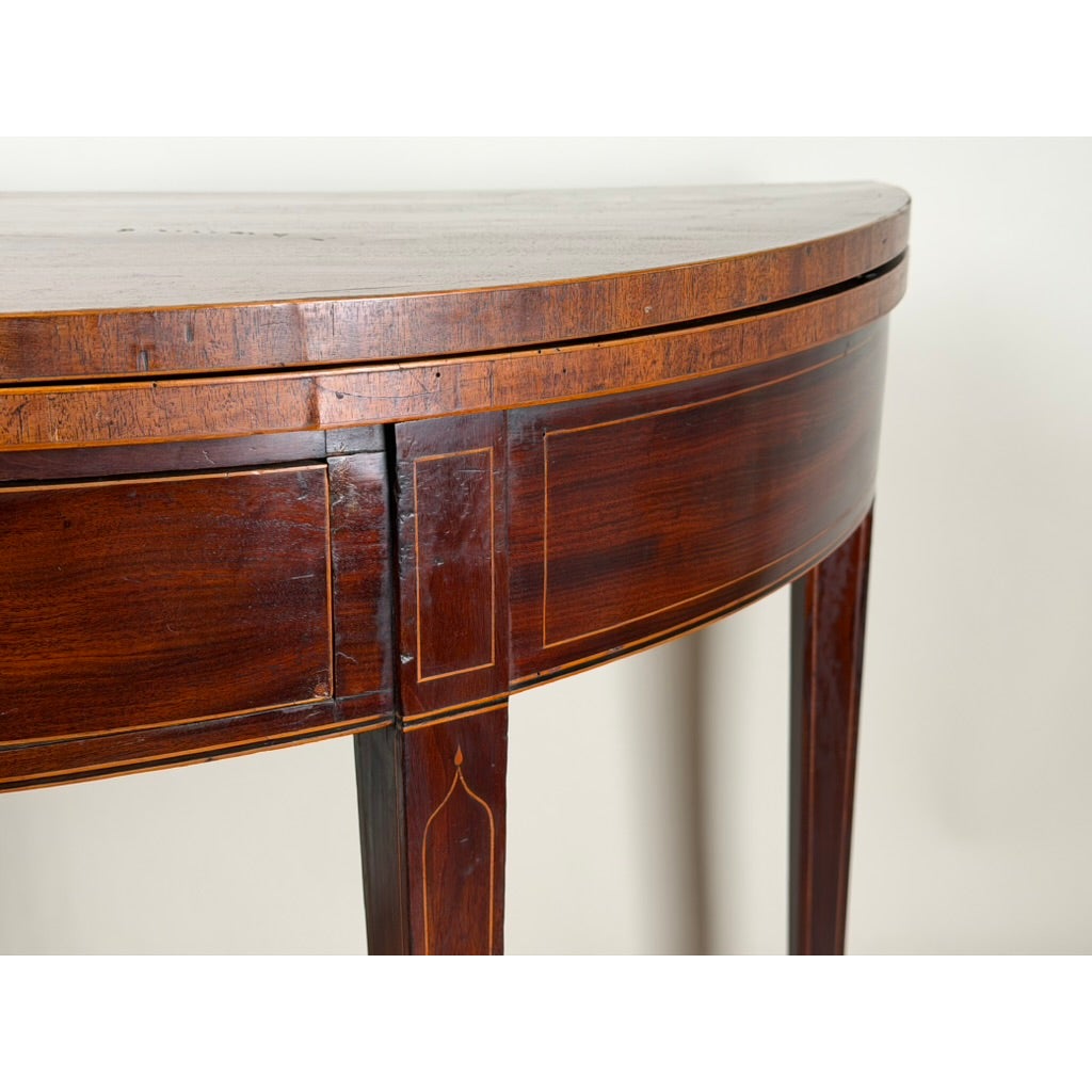 An English George III Mahogany and Satinwood-Inlaid Demilune Games Table, circa 1790–1810, featuring a Hepplewhite-style semicircular shape with inlaid light-colored trim and a tapered leg.