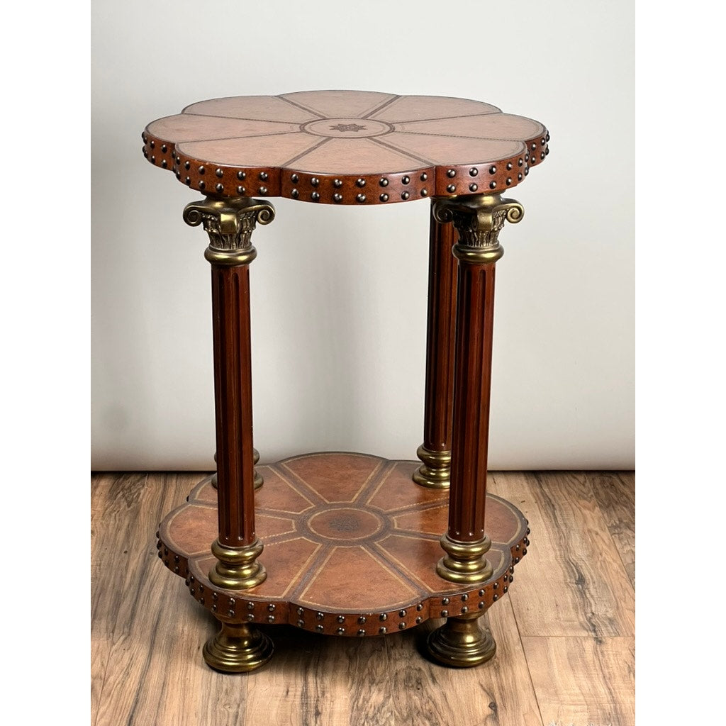 The Vintage Maitland-Smith Leather-Clad Occasional Table by Maitland Smith features a round scalloped top, lower shelf, brass nailhead trim, and three fluted columns with gold-toned accents. This elegant piece enhances any wood floor setting.