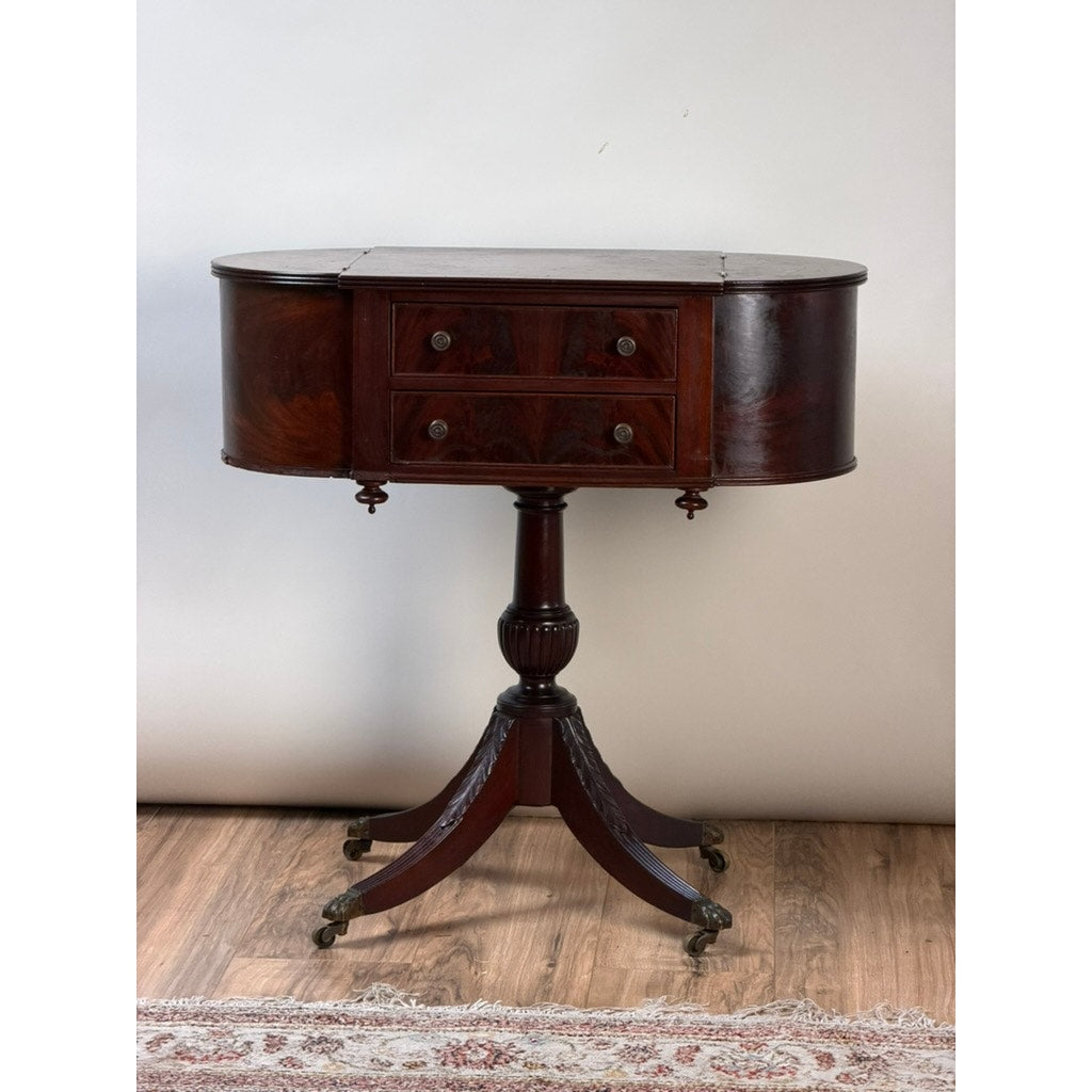 Early 19th Century New York Classical mahogany astragal-end work table in the manner of Duncan Phyfe, c. 1815–1825, featuring curved sides, four drawers, a central pedestal, and four splayed legs on brass castors—displayed on a wooden floor with part of a rug visible. This piece exemplifies the refined elegance and craftsmanship characteristic of early 19th-century parlor furnishings. Brand: None/Unknown.