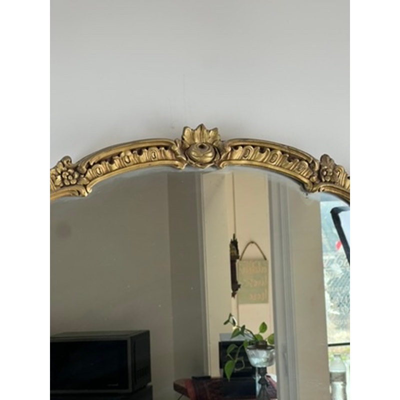 Antique Gilt Wood Oval Mirror (early 20th century) with floral details reflects part of a room, showing a plant, wall art, microwave, and a sunlit window. Brand unknown.