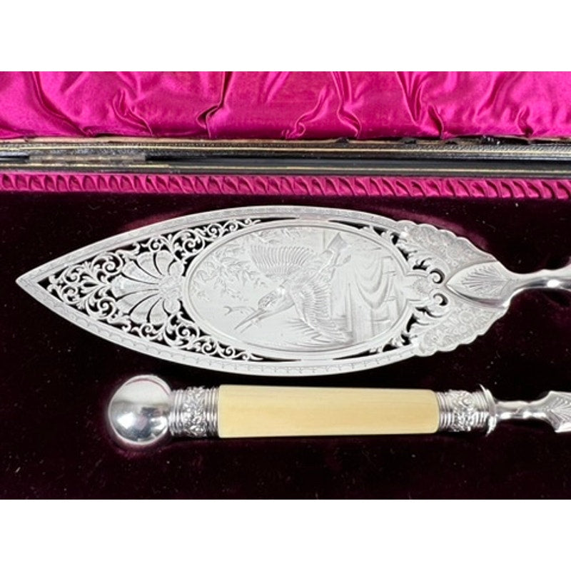 A close-up of an ornate silver-plated fish slice with detailed engravings and a cream handle, displayed on velvet with pink satin—part of an Antique Victorian Silver-Plated Fish Serving Set, complete with serving knife and fork.