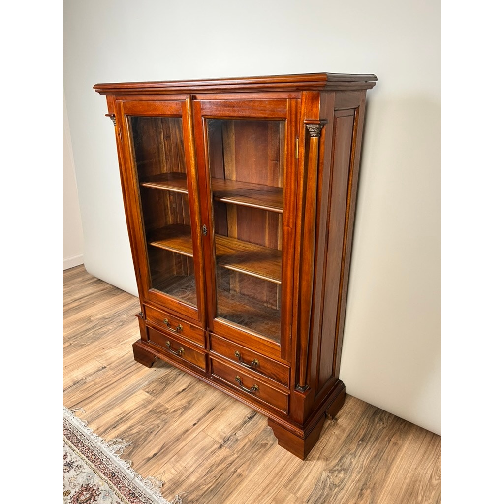 The Vintage Victorian Style Glass Door Mahogany Bookcase features two glass doors, three interior shelves, and three lower drawers with decorative brass handles. Its polished finish adds elegance as it stands on a wood floor against a plain wall.