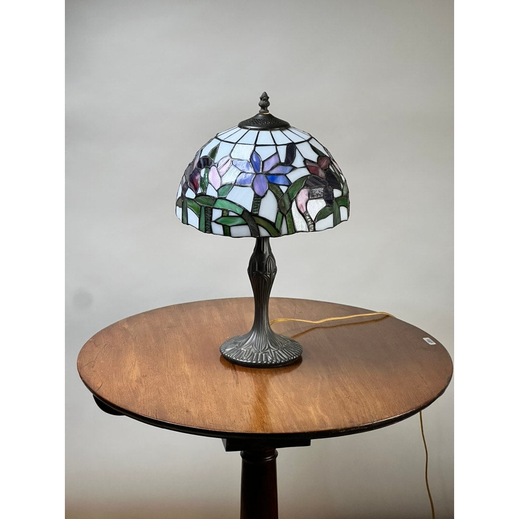 A Vintage Tiffany Style Floral Stained Glass Table Lamp with a decorative metal base sits on a round wooden table against a plain background, its cord visible on the tabletop. Brand: None and/or Unknown.