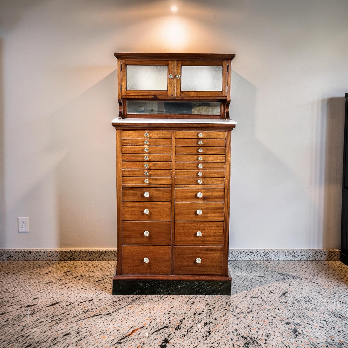 An Antique Mahogany Multi-drawer Dental Cabinet by The American Cabinet Co. (early 20th c.), featuring white round knobs, marble slab, and frosted glass doors, stands on a polished stone floor against a light-colored wall.