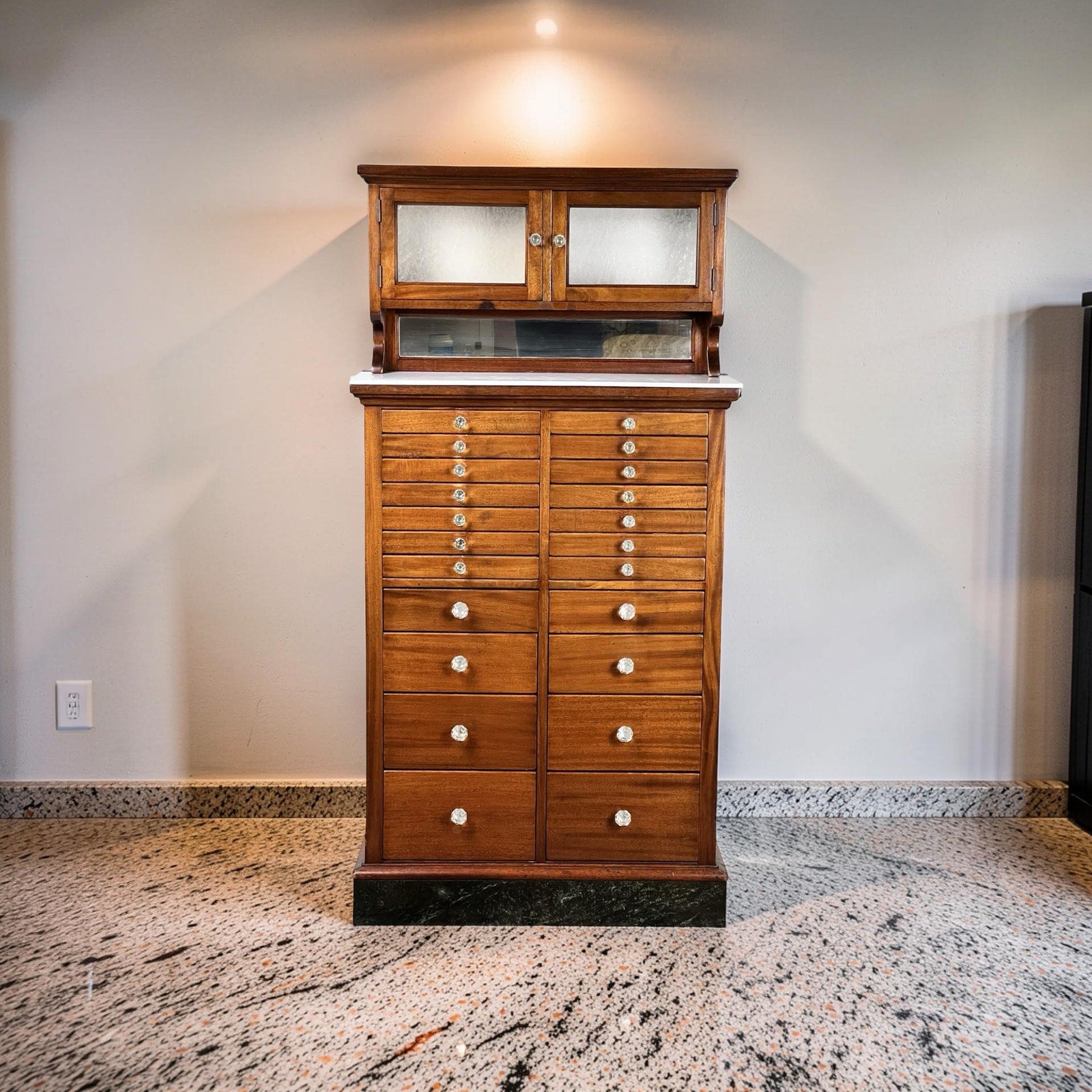 An Antique Mahogany Multi-drawer Dental Cabinet by The American Cabinet Co. (early 20th c.), featuring white round knobs, marble slab, and frosted glass doors, stands on a polished stone floor against a light-colored wall.