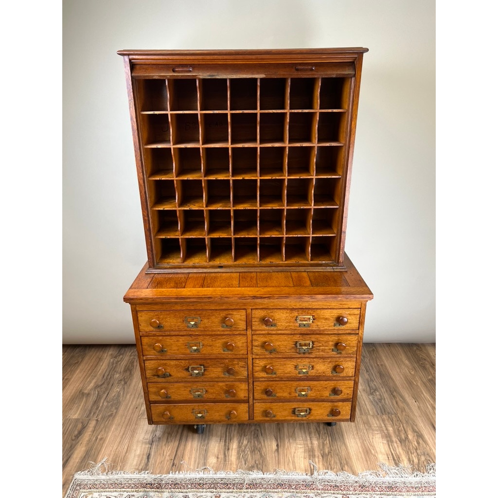 An Antique Early 20th Century Oak Multidrawer File with Roll Top Pigeon Hole Top stands on a wooden floor, part of a rug visible at the bottom. Brand: None and/or Unknown.