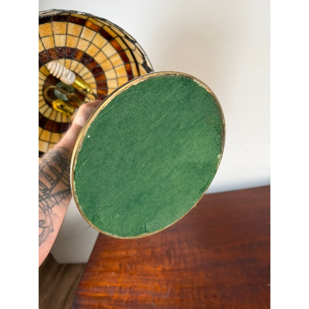 A hand holds the Vintage Tiffany Revival Leaded Stained Glass Table Lamp with a round green felt bottom and a cast brass tree-form base from Pearl City Antiques, revealing part of a tattooed arm; a wooden table and white wall appear in the background.