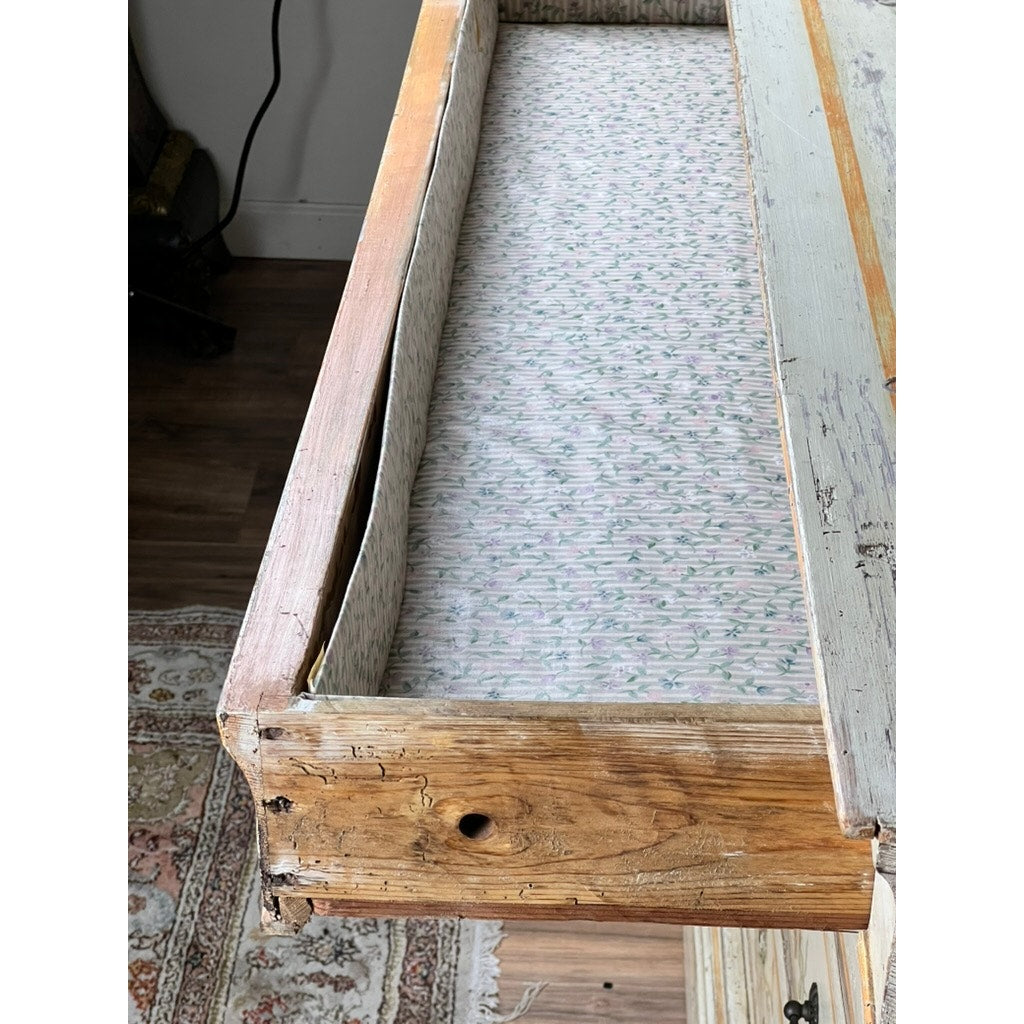 An open drawer from an Antique Italian Paint Decorated Four Drawer Commode (Early 19th Century) reveals a floral liner and worn front, placed in a rustic room with a patterned rug and wood floor. Brand: Unknown.