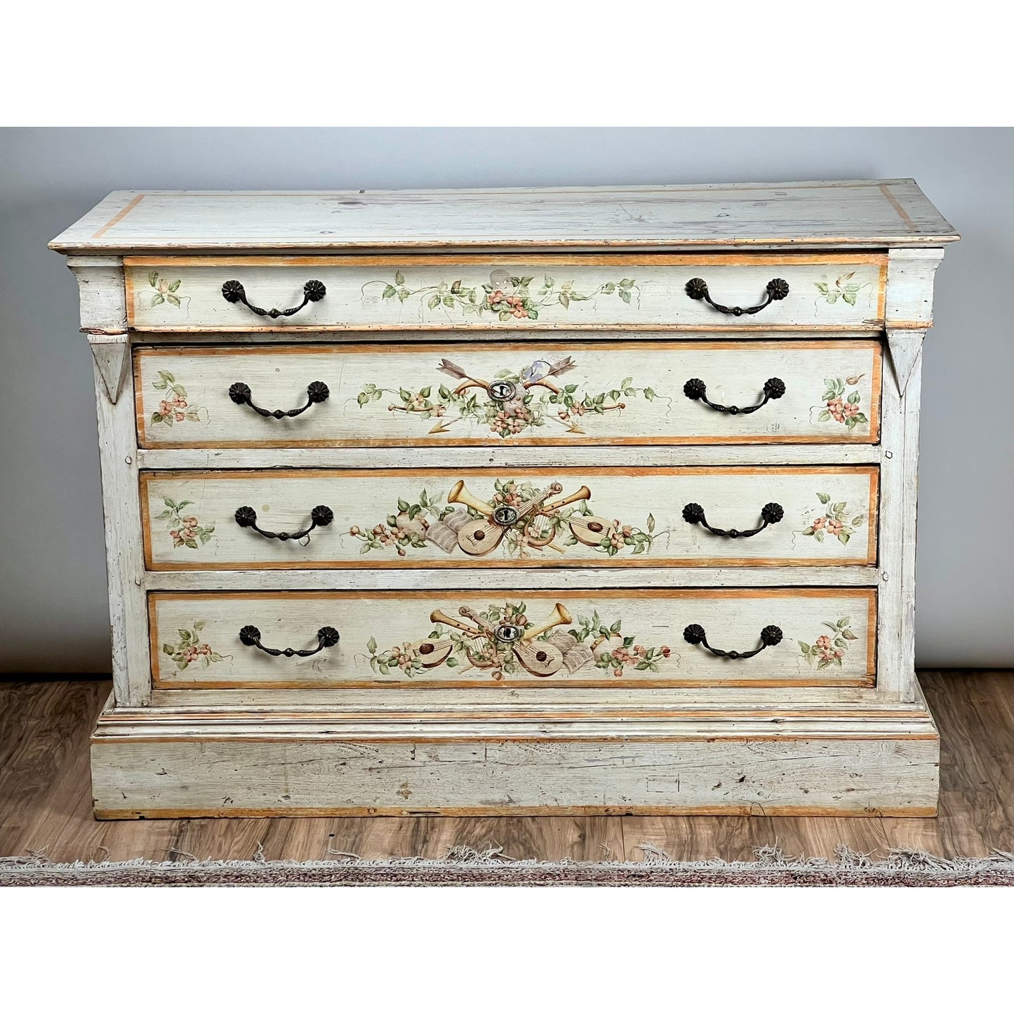 An Antique Italian Paint Decorated Four Drawer Commode (early 19th century) features ornate hand-painted floral and musical designs with black metal handles, set on a wooden floor against a plain gray wall. Brand: Unknown.