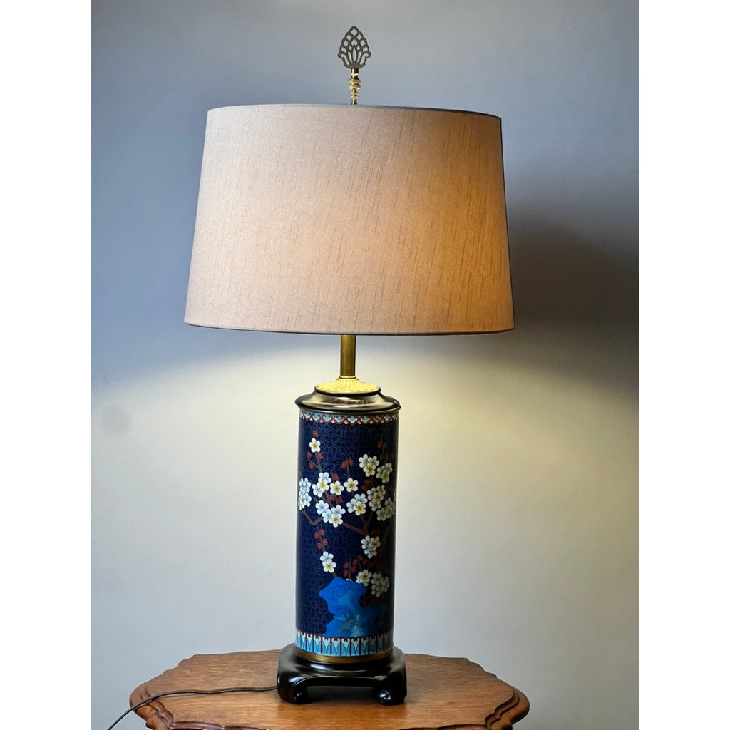 A Vintage Japanese Cloisonné Cylinder Table Lamp by Knob Creek, featuring a beige shade and dark floral-patterned ceramic base, sits on a wooden table against a light gray wall, casting a warm classic glow.