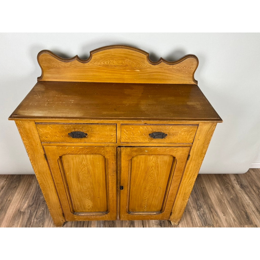An Antique Victorian Grain Painted Country Jelly Cupboard c. 1870 stands against a light-colored wall on a wood floor, featuring two drawers, cabinet doors, a decorative backsplash, and dark metal handles. Brand: None/Unknown.