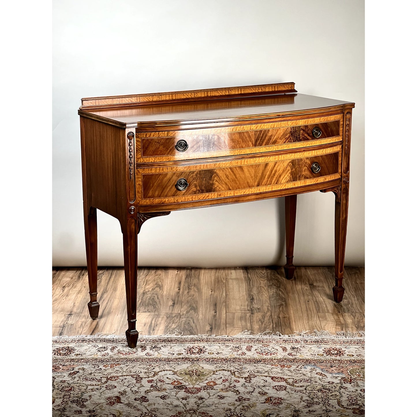 The Vintage Federal Style Bow Front Mahogany Sideboard by Paine Furniture Boston features two drawers, ornate metal handles, and tapered legs. This timeless Pa piece stands on a wooden floor beside a patterned rug against a light-colored wall.