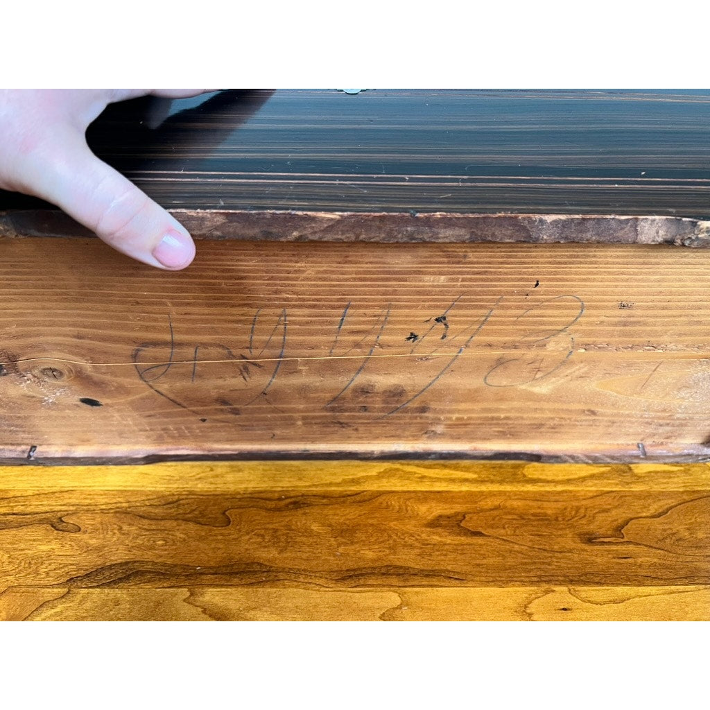 A hand lifts the edge of an Antique Swiss 12 Tune Music Box (c. Late 19th Century), revealing faded black writing inside the wood; the surface shows natural grain patterns and color variations. Brand unknown.