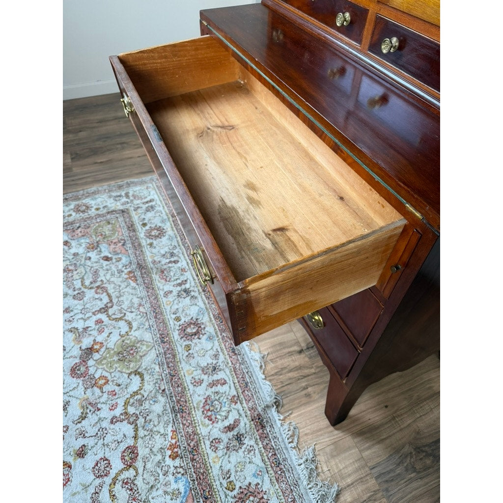 An open wooden drawer in the Federal Flame Mahogany Secretary Bookcase with Eagle Finial, c. 1810–1825, reveals its empty interior. The bookcase, crafted in rich flame mahogany and adorned with brass handles, exemplifies the refined style of early 19th-century American cabinetmaking and features additional drawers above and below. Beside it lies a patterned area rug on a wood floor.