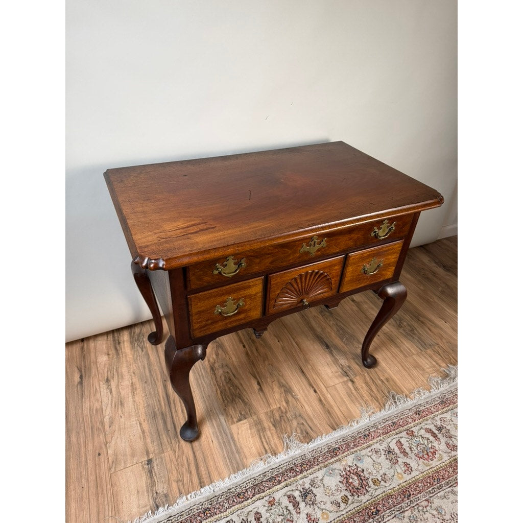 An 18th Century American Queen Anne Walnut Shell-Carved Lowboy (c. 1740–1770), featuring claw-foot legs, brass handles, three drawers, and an ornate carved shell detail—evoking classic antique elegance—set on a wood floor beside a patterned rug.