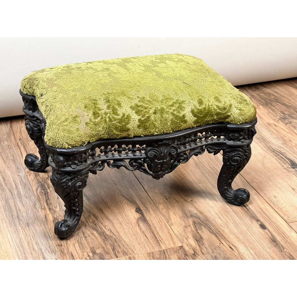 The Antique Victorian Cast Iron Foot Stool c. 1900 features a green patterned upholstered cushion and cast iron legs, showcasing antique elegance, shown on a wooden floor against a plain white background. Brand: None/Unknown.