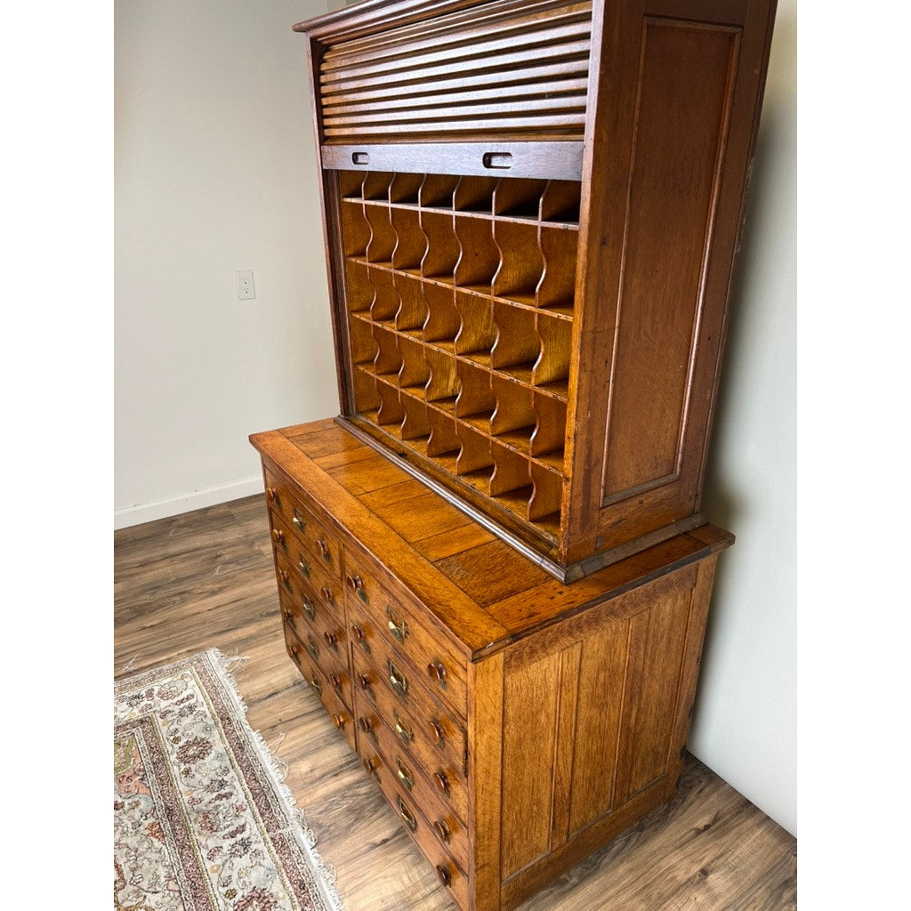 An Antique Early 20th Century Oak Multidrawer File with Roll Top Pigeon Hole Top stands against a white wall on a wooden floor, with part of a patterned rug visible. Brand: None and/or Unknown.