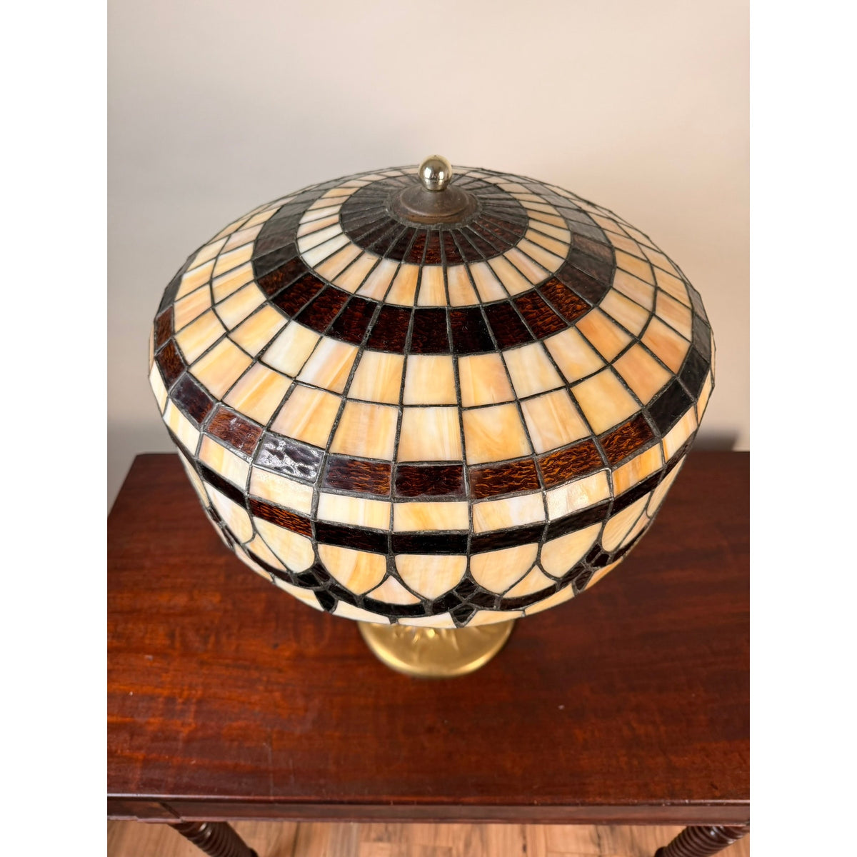 The Pearl City Antiques Vintage Tiffany Revival Leaded Stained Glass Table Lamp with Cast Brass Tree-Form Base features a geometric stained glass shade in cream, brown, and black, gracefully displayed on a wooden table. The artistic cast brass tree-form base beautifully enhances the classic Tiffany-style aesthetic.