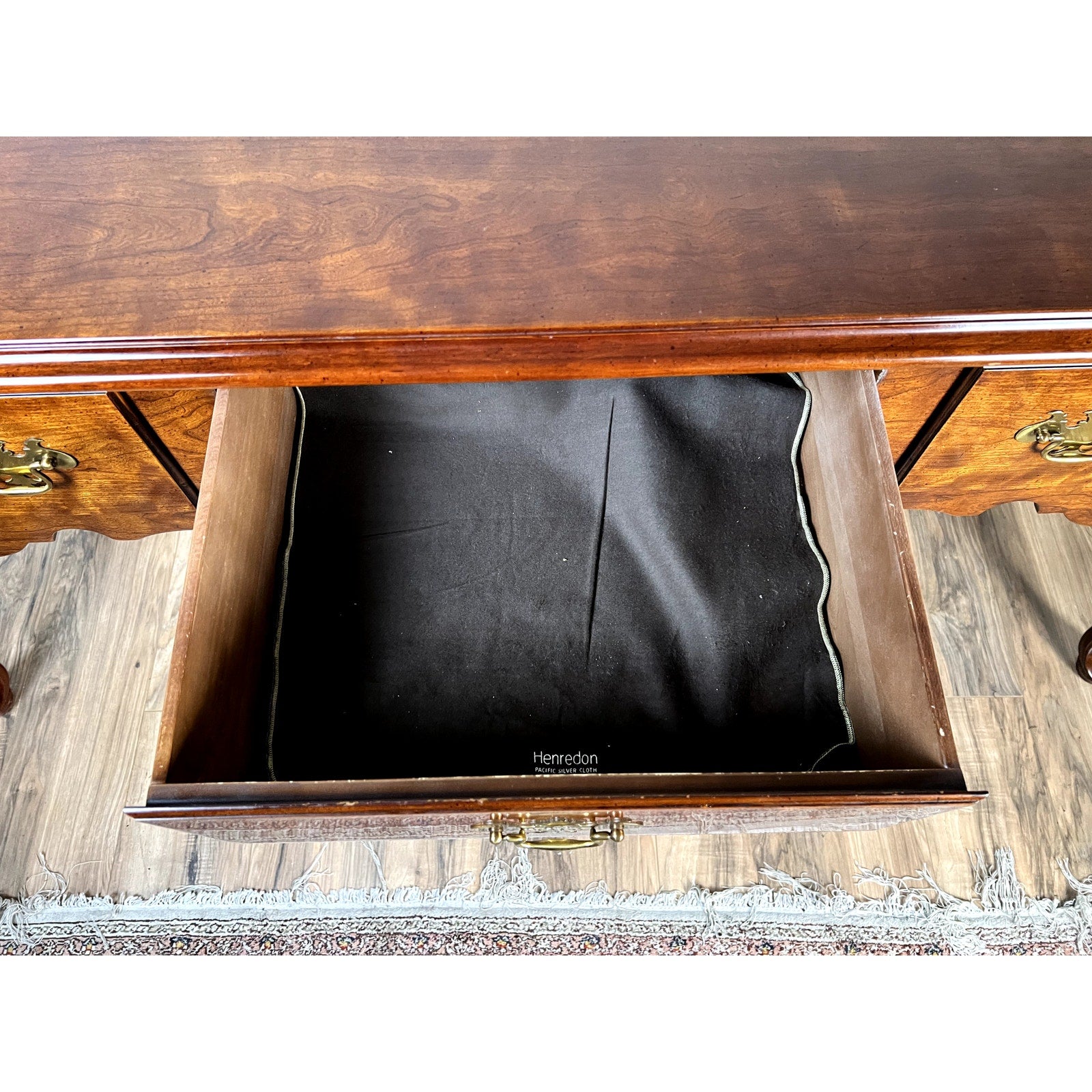 An open wooden drawer lined with black fabric inside a Vintage Solid Cherry Queen Anne Style Sideboard by Henredon, featuring brass handles and part of a rug on a wood floor.