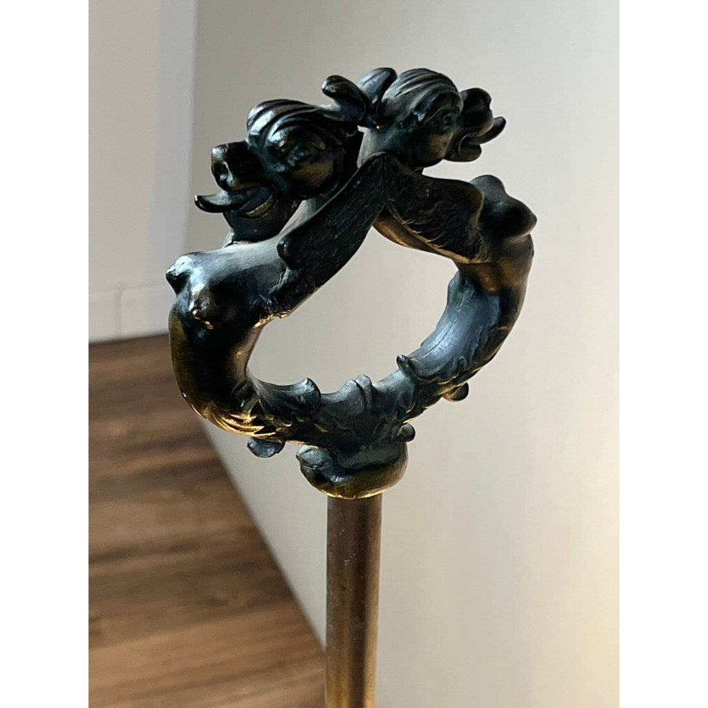 The Antique Five Burner Lucerne Whale Oil Lamp with Napoleon I Fireplace Screen Shade features a bronze handle with two winged, nude female figures with flowing hair back-to-back in a circular form, set against a wooden floor and plain wall.