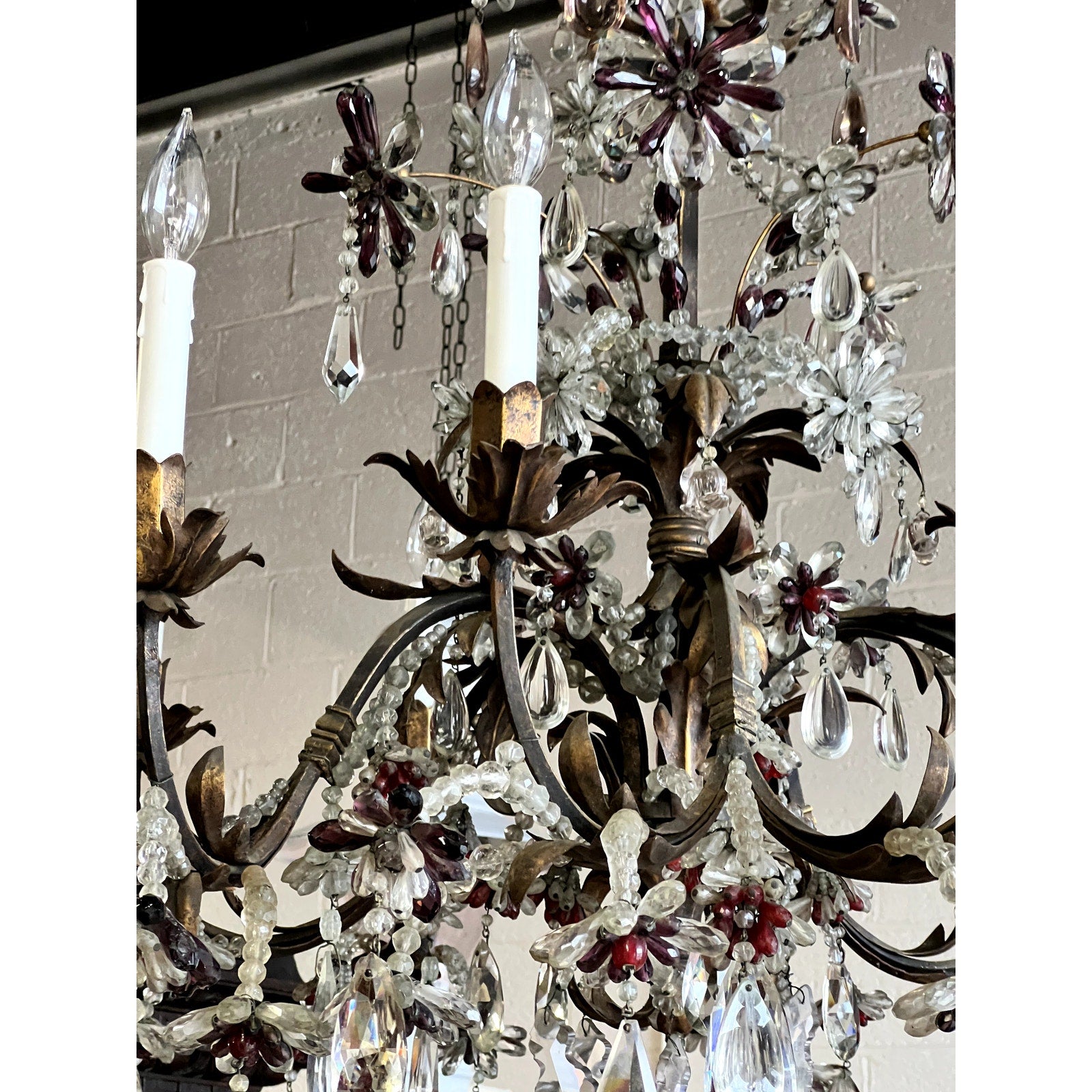 A detailed view of an Antique Late 19th Century French Rococo Tole Amethyst and Crystal Ten Light Chandelier, featuring ornate metal scrollwork, floral-shaped amethyst and clear prisms, and faux candle lights against a light brick wall.