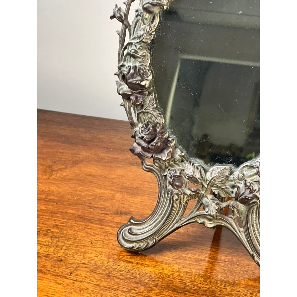 Close-up of the Antique Art Nouveau Silvered Cast Brass Rose Vanity Dressing Mirror, dated 1909, with an intricate floral and leaf design on its metal frame, resting on a wooden surface. The decorative stand and mirror edge are visible.