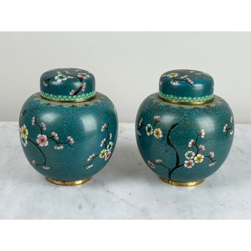 A vintage pair of Chinese cloisonné ginger jars, featuring teal ceramic with pink and yellow flower branches, gold-colored bases and rims, and lids, are displayed side by side on a light marble surface. Brand: Unknown.