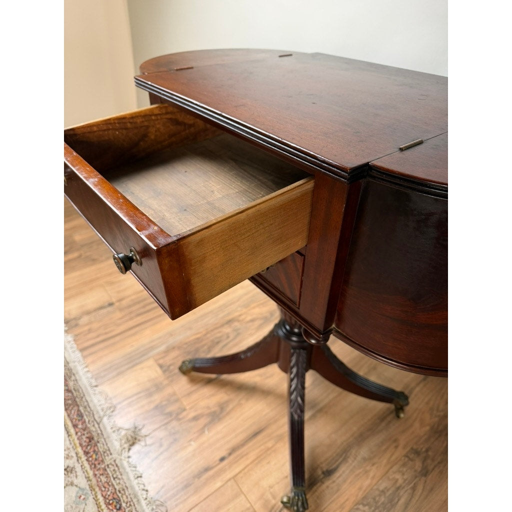 An early 19th century New York Classical mahogany astragal-end work table in the manner of Duncan Phyfe (c. 1815–1825), featuring an open drawer and standing on a carved pedestal base with four legs, brings historical refinement to the space. Set against a cream-colored wall and positioned on a wooden floor near a rug, this piece recalls the elegance of parlor furnishings from its era.