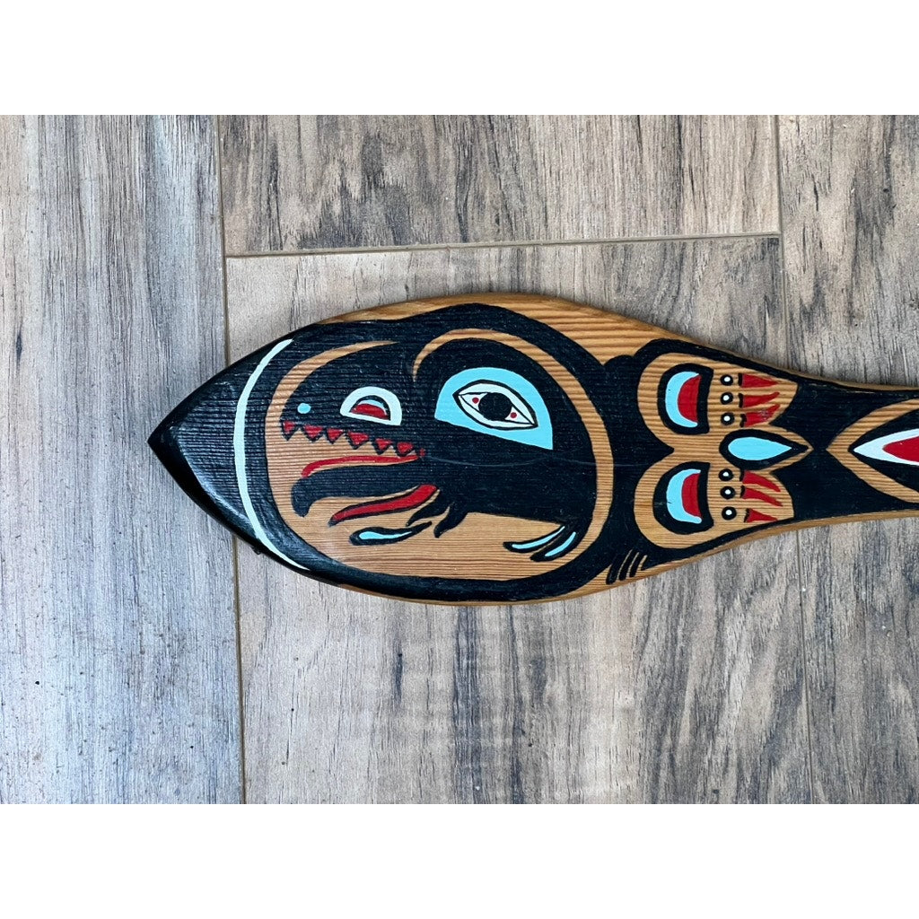 A Vintage Inuit Carved Red Cedar Paddle from Sitka, Alaska (signed illegibly) with a traditional animal motif in bold black, red, and turquoise rests blade-down on a wood floor. Only the ceremonial paddle’s blade is visible.