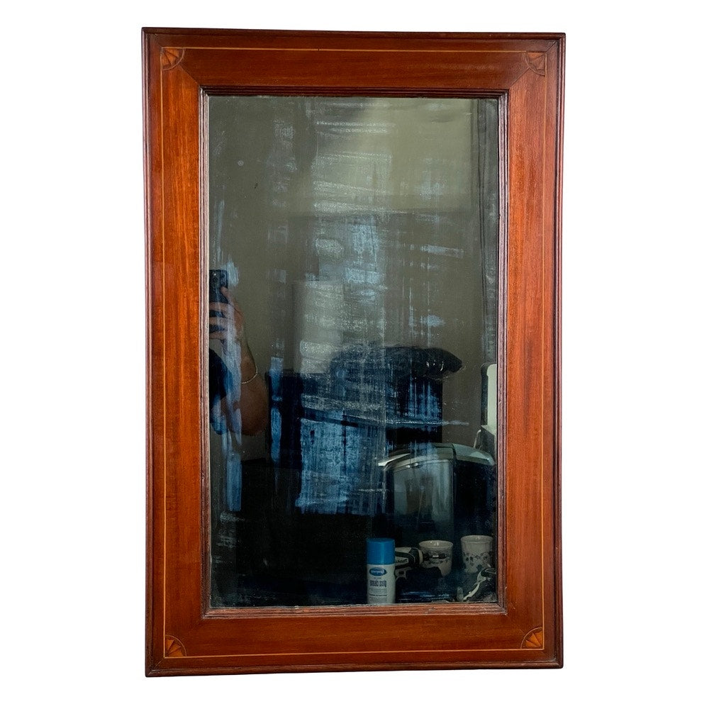 An American Federal mahogany wall mirror with fan inlay, featuring its original hand-silvered glass (circa 1810–1825). The antique mirror's wooden frame surrounds a surface marked with visible streaks and smudges that speak to its history. In the reflection, a softly blurred room, a person taking a photo, and several objects near the bottom edge are visible. Brand: None/Unknown.
