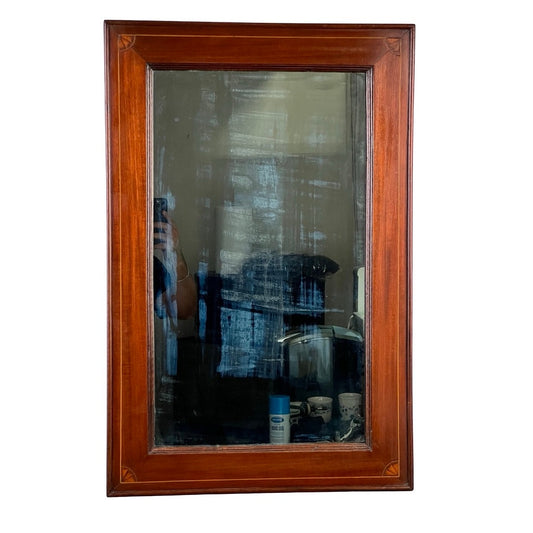 An American Federal mahogany wall mirror with fan inlay, featuring its original hand-silvered glass (circa 1810–1825). The antique mirror's wooden frame surrounds a surface marked with visible streaks and smudges that speak to its history. In the reflection, a softly blurred room, a person taking a photo, and several objects near the bottom edge are visible. Brand: None/Unknown.